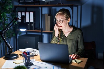 Young blonde woman working at the office at night looking stressed and nervous with hands on mouth...