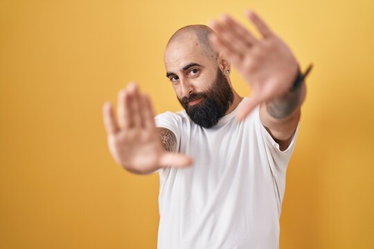 Young hispanic man with beard and tattoos standing over yellow background doing frame using hands palms and fingers, camera perspective