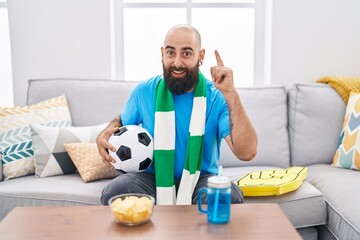 Young hispanic man with beard and tattoos football hooligan holding ball supporting team surprised with an idea or question pointing finger with happy face, number one