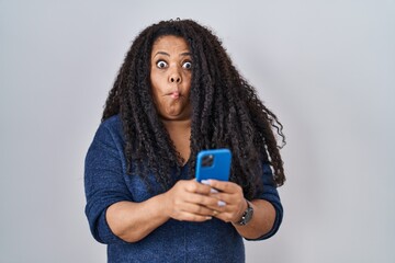 Plus size hispanic woman using smartphone typing message making fish face with mouth and squinting...