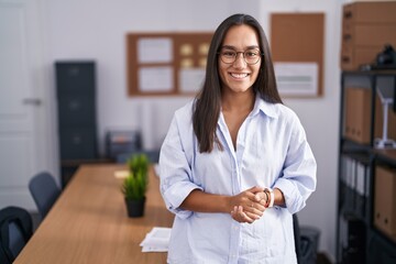 Young hispanic woman at the office with hands together and crossed fingers smiling relaxed and cheerful. success and optimistic