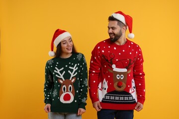 Happy young couple in Santa hats showing Christmas sweaters on orange background