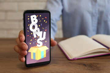 Bonus gaining. Woman showing smartphone at wooden table, closeup. Illustration of open gift box, word and confetti on device screen