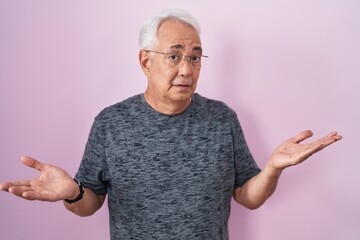 Middle age man with grey hair standing over pink background clueless and confused expression with...