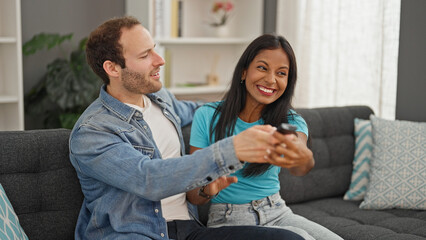 Beautiful couple sitting on sofa fighting for tv remote control at home