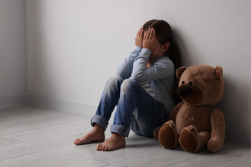 Child abuse. Upset little girl with teddy bear sitting on floor near light wall indoors, space for...