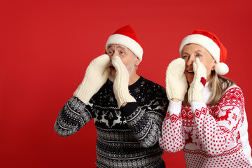 Senior couple in Christmas sweaters, Santa hats and knitted mittens shouting something on red background
