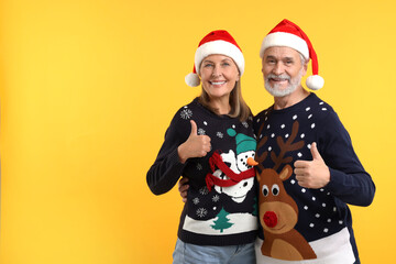 Fototapeta premium Happy senior couple in Christmas sweaters and Santa hats showing thumbs up on orange background. Space for text