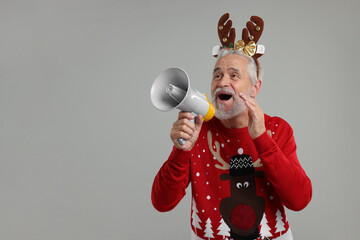 Senior man in Christmas sweater and reindeer headband shouting in megaphone on grey background. Space for text