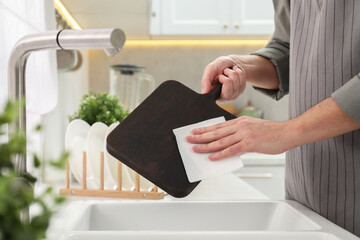 Man wiping dark wooden cutting board with paper napkin at sink in kitchen, closeup