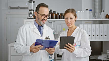 Two scientists engrossed in reading an intriguing document on clipboard at the lab, speaking about their startling discovery, while working on a touchpad together.