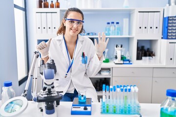 Young hispanic woman working at scientist laboratory showing and pointing up with fingers number...