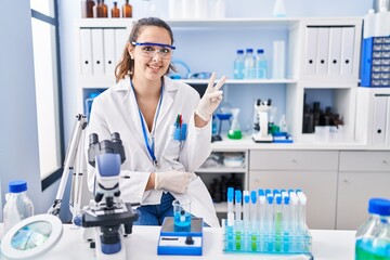 Young hispanic woman working at scientist laboratory smiling with happy face winking at the camera doing victory sign. number two.