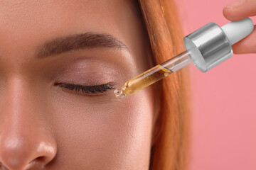 Obraz premium Beautiful young woman applying cosmetic serum onto her face on pink background, closeup