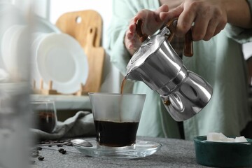 Woman pouring aromatic coffee from moka pot into glass at table in kitchen, closeup