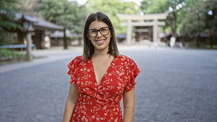 Cheerful, beautiful hispanic woman with glasses poses confidently, smiling at tokyo's meiji shrine, her joy unequivocally radiating through her astonishing smile