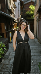 Confident hispanic woman, beautiful and joyful, cheerfully posing with glasses on, standing and smiling on kyoto's traditional streets, radiating natural and carefree latin expression.