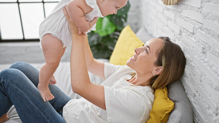 Joyful mother lying on bed, confidently holding happy baby daughter mid-air, having fun while smiling and playing together in warm, homely bedroom.