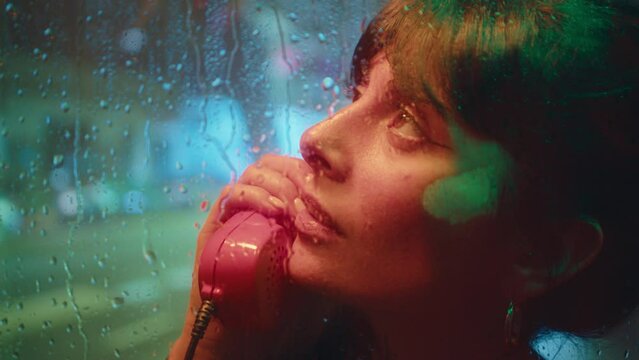 Young beautiful woman standing behind wet glass in phone booth and talking on payphone at rainy night in the city