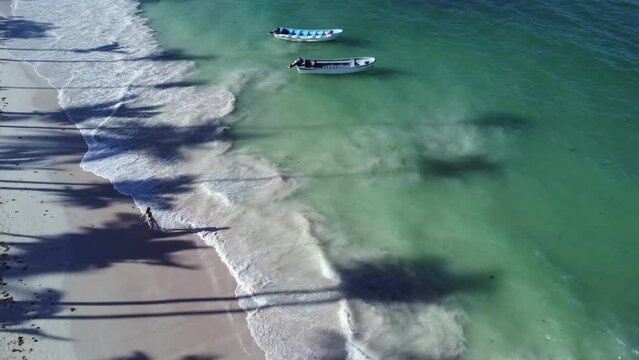 Ujęcie z drona na wesołą kobietę biegnącą do turkusowego oceanu, a obok stoją dwie łódki (motorówki). 