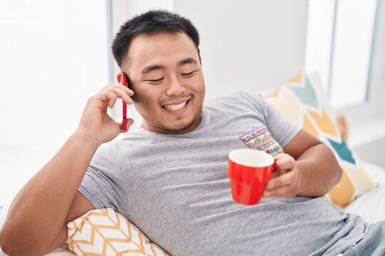 Young chinese man talking on smartphone drinking coffee at bedroom