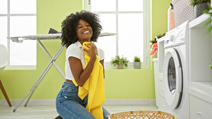 African american woman washing clothes holding clean sweater smiling at laundry room - obrazy, fototapety, plakaty