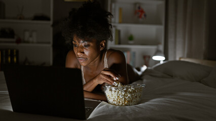 African american woman wearing lingerie watching movie on laptop eating popcorn at bedroom