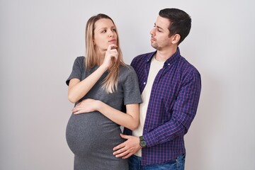 Young couple expecting a baby standing over white background with hand on chin thinking about question, pensive expression. smiling and thoughtful face. doubt concept.