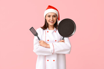 Beautiful female chef in Santa hat with frying pan and spatula on pink background