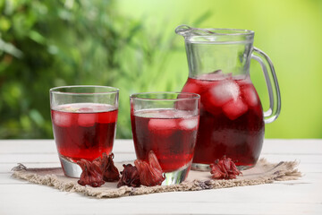 Refreshing hibiscus tea with ice cubes and roselle flowers on white wooden table against blurred green background