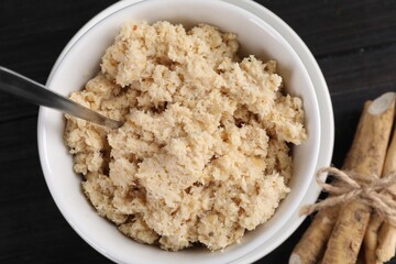 Bowl of tasty prepared horseradish, spoon and roots on black wooden table, flat lay