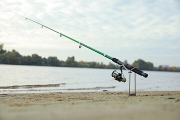 Fishing rod with reel on sand near river, space for text