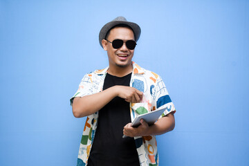 Smiling Asian tourist man holding digital tablet and pointing finger and looking at camera isolated on light blue background. vacation concept.