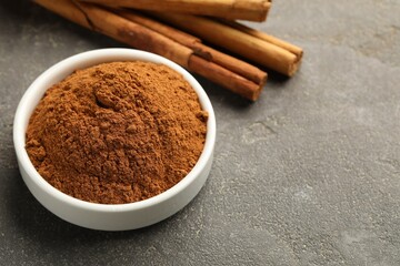 Bowl of cinnamon powder and sticks on grey table, closeup. Space for text