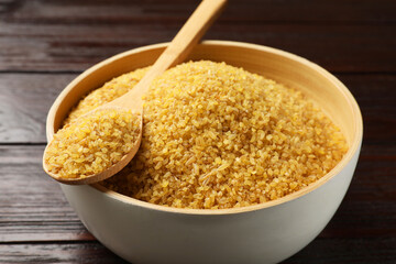 Bowl and spoon with raw bulgur on wooden table, closeup