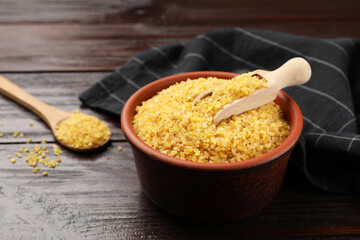 Bowl and spoon with raw bulgur on wooden table, closeup