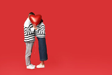 Loving young couple hiding behind heart-shaped balloon on red background. Celebration of Saint...
