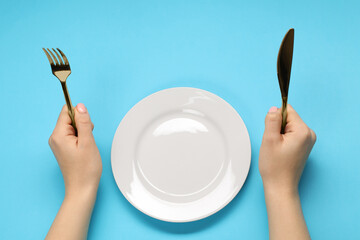 Woman holding fork and knife near empty plate at light blue table, above view