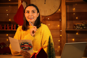 Happy young woman writing Christmas greeting card at table in festive room