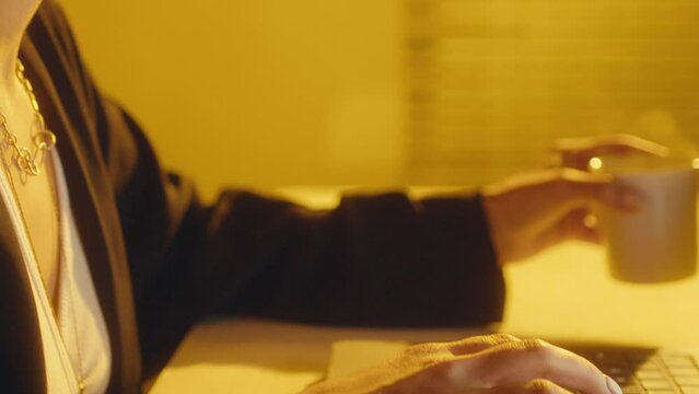 Woman typing on laptop keyboard and drinking hot tea or coffee from mug at desk in room with bright yellow light. Close-up view, tilt up shot