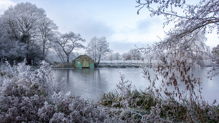 A view across a frozen pool towards an old corrugated boat house. Taken in a heavy frost, the vegetation and grasses are white over. The water is frozen - 688328132