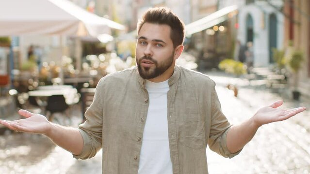I dont know what to say. Confused young man feeling embarrassed about ambiguous question, having doubts, no idea, being clueless uncertain outdoors. Caucasian adult guy standing in urban city street