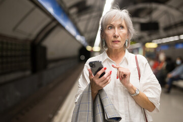 Senior woman with cell phone in hand standing in subway station near railway and waiting for train.