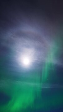 A view towards the sky in the arctic night. The moon is surrounded by a  halo and light clouds as an aurora is dancing elegantly beneath.