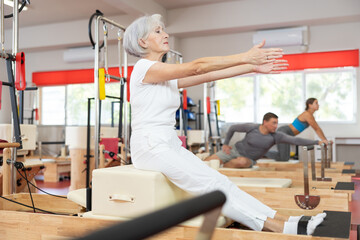 Fototapeta na wymiar Elderly athletic woman doing stretching on pilates machine in pilates studio..