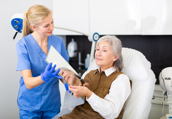 Fototapeta premium Senior woman looking in mirror after cosmetological procedures. Female cosmetologist standing near and looking at her.