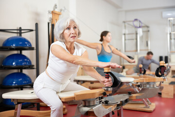 In gym, senior female performs Pilates exercises to lose weight and bring muscles of arms, chest, and body into tone using heron pulley tower. Comprehensive equipment for pumping whole body