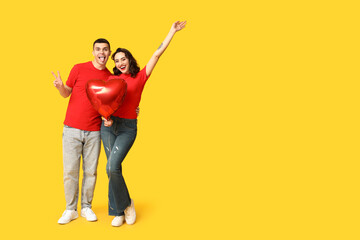 Loving young couple with heart-shaped balloon showing victory gesture on yellow background. Celebration of Saint Valentine's Day
