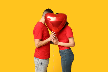 Loving young couple hiding behind heart-shaped balloon on yellow background. Celebration of Saint...