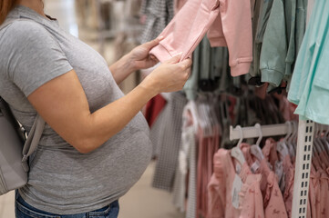 Caucasian pregnant woman chooses baby clothes in a store. Faceless expectant mother in the 3rd trimester. 